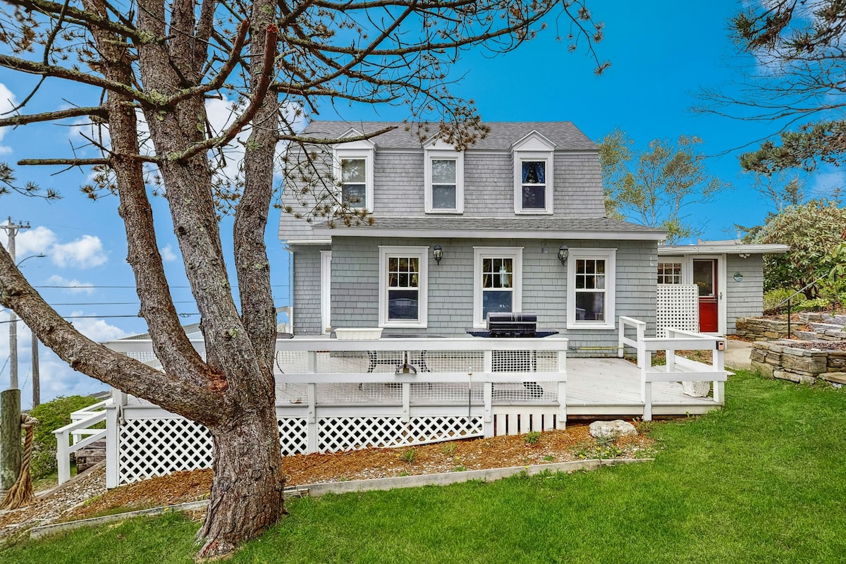 The exterior of a classic gray cottage is visible, surrounded by a manicured lawn. A large deck with a dining area faces the water, while a tree offers shade. The cottage features multiple windows and a red door, contributing to its charming character.