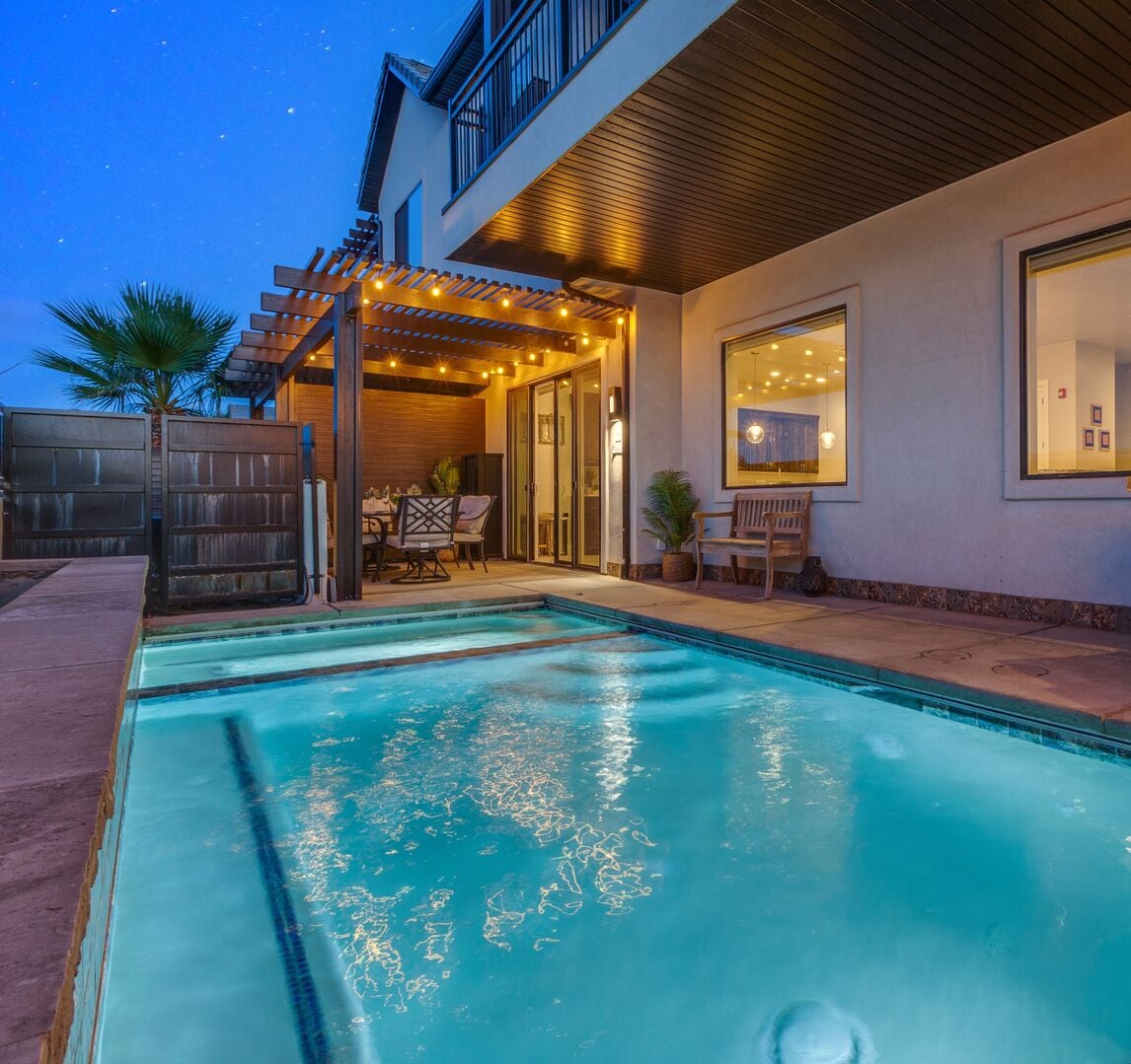 A private pool is surrounded by a spacious patio, featuring a covered seating area with outdoor furniture. Soft lighting illuminates the area, creating a serene atmosphere. Palm trees are positioned nearby, contributing to the tranquil setting under the evening sky.