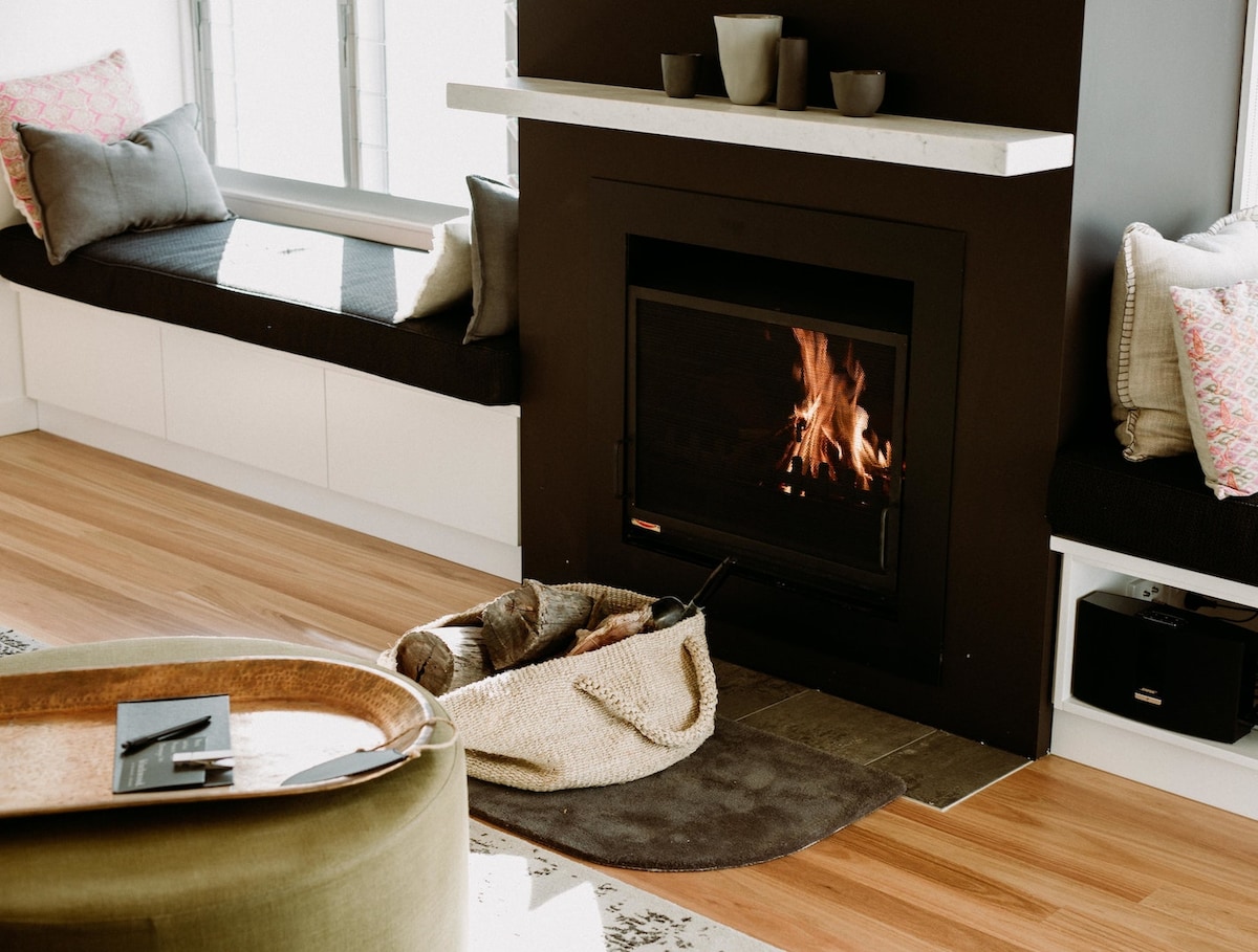A modern fireplace is set within a sleek black feature wall, surrounded by comfortable seating areas with plush cushions. A stylish basket holds logs nearby, and natural light fills the space through expansive windows, enhancing a cozy ambiance.