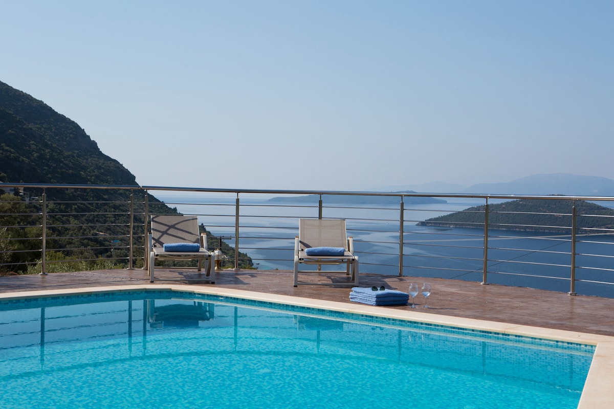 A private swimming pool glimmers in the sunlight, surrounded by a sleek railing. Two lounge chairs with blue cushions are positioned nearby, offering serene views of the Ionian Sea and rolling hills in the distance, all under a clear, bright sky.