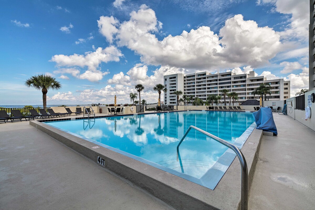 A heated pool is surrounded by lounge chairs, with palm trees lining the edge. The clear water reflects a vibrant sky filled with clouds. The condo complex is visible in the background, contributing to the inviting atmosphere.