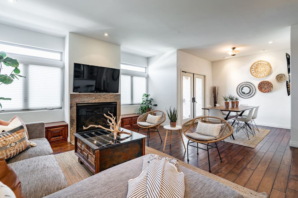 A living area features a modern fireplace paired with stylish seating arranged around a coffee table. Large windows provide natural light, while a dining space with a circular table is visible in the background. Decorative wall art adds subtle character to the room.