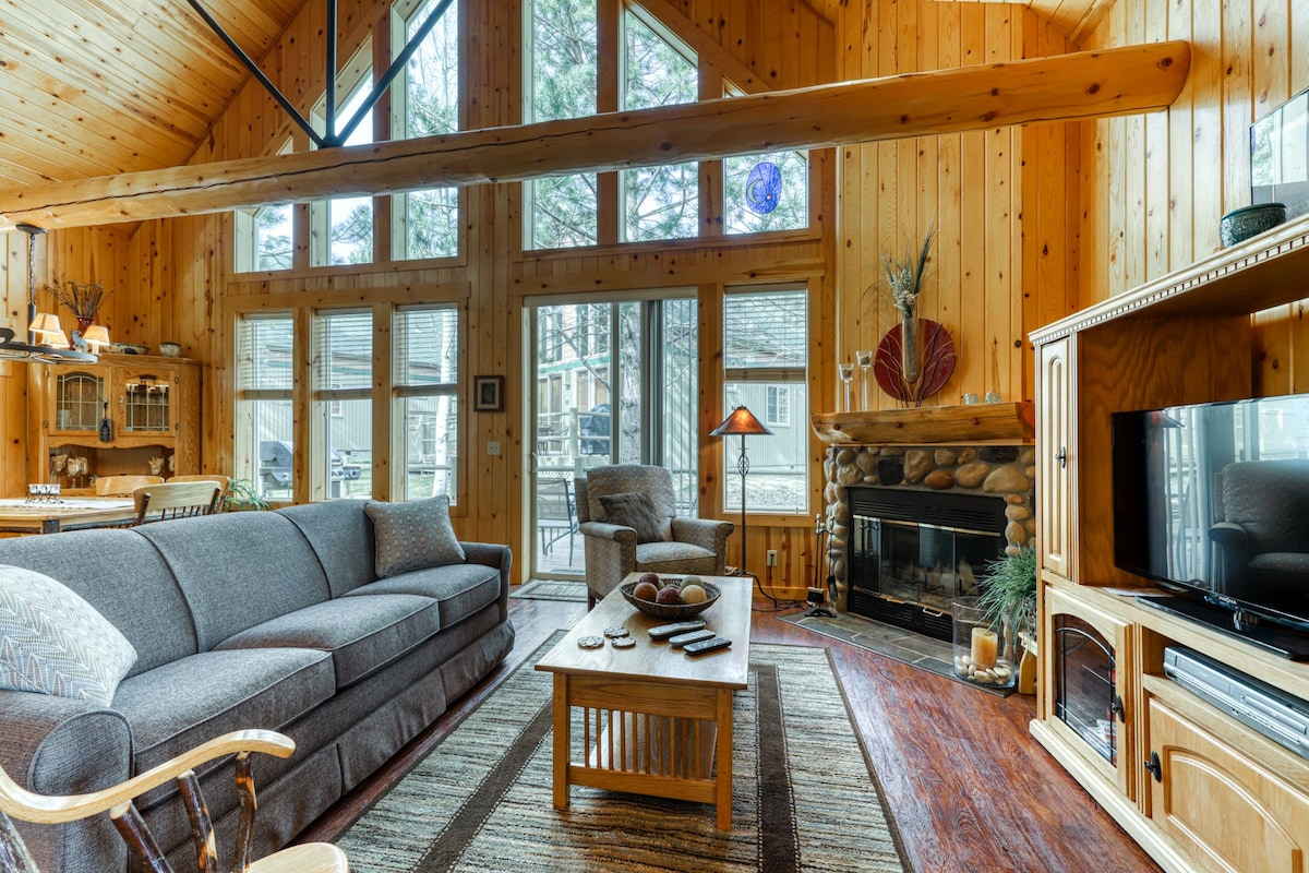 A spacious living area features vaulted pine ceilings and large windows that illuminate the rustic wood interior. A comfortable gray sofa is positioned near a wooden coffee table and a stone fireplace, creating a cozy focal point. A seating area with a chair is visible beside the patio doors.