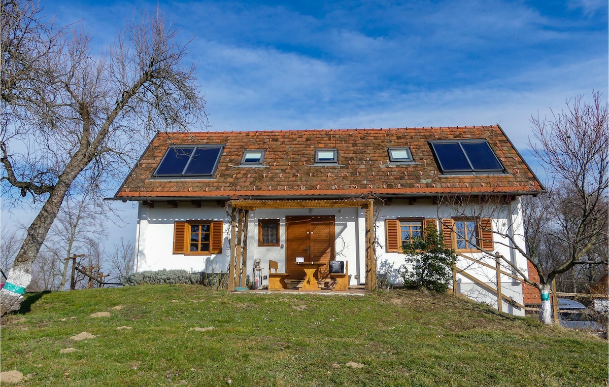 The exterior of the Kellerstöckl showcases a two-story structure with a sloped roof featuring solar panels. A wooden entrance is highlighted by a pergola, surrounded by greenery. Large windows invite natural light, and a tranquil scene of rolling hills complements the inviting presence of the house.