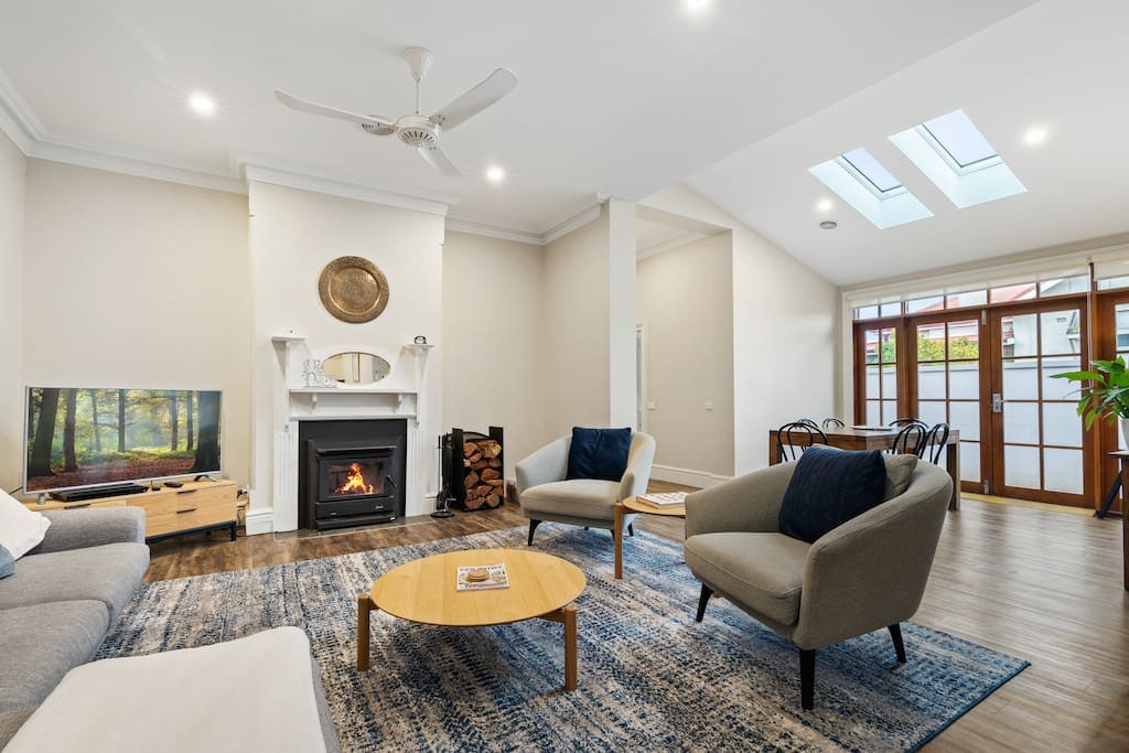 A cozy living area includes two comfortable armchairs and a spacious sofa, arranged around a circular coffee table. A TV is mounted on a light wooden unit, while a hearth is bordered by stacked firewood. Natural light enters through skylights and glass doors.