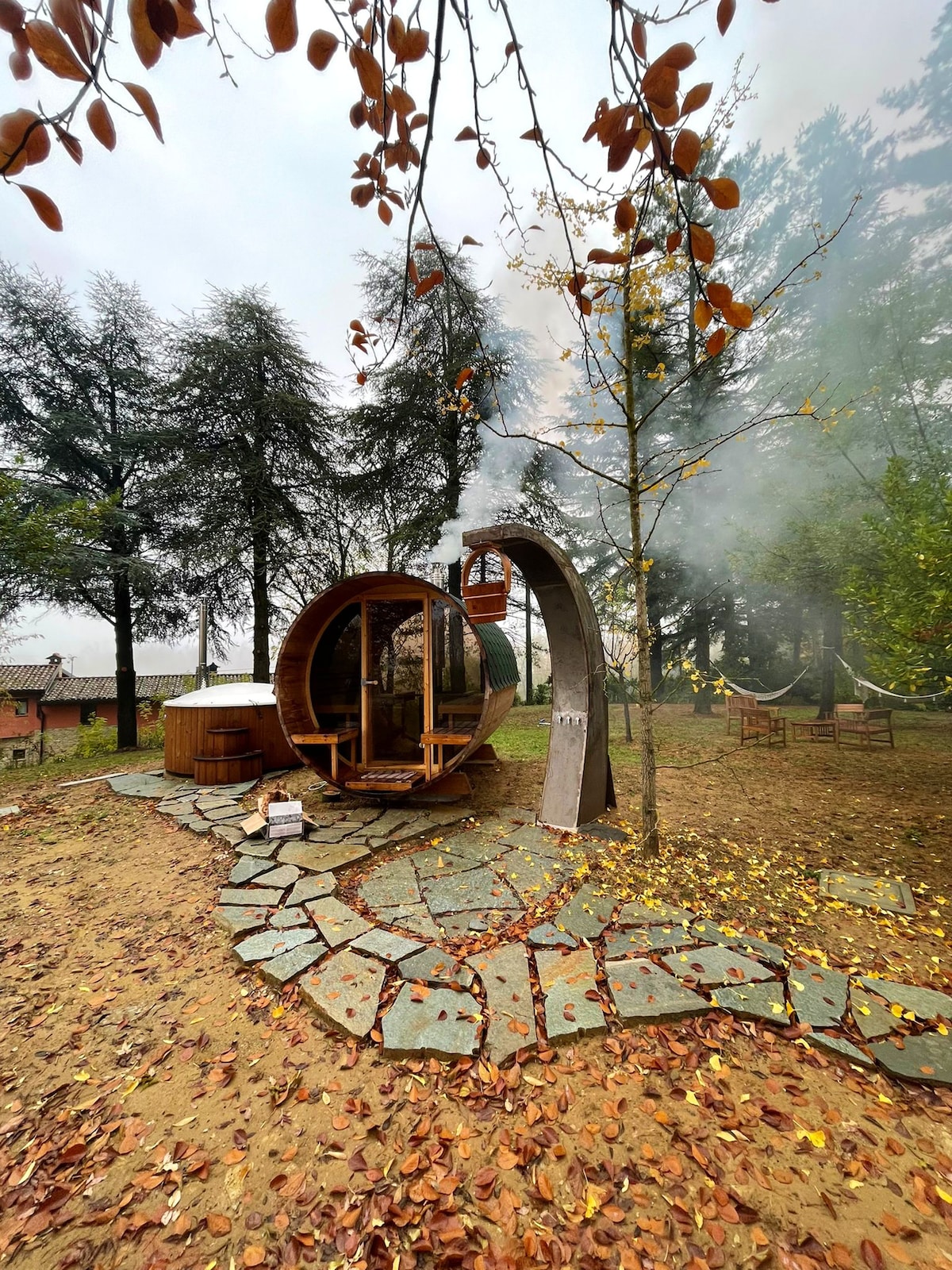 An outdoor relaxation area features a wooden sauna and hot tub surrounded by nature, with a stone path leading to them. Smoke gently rises from the sauna chimney, and seating options are placed nearby, creating a serene atmosphere among the trees.