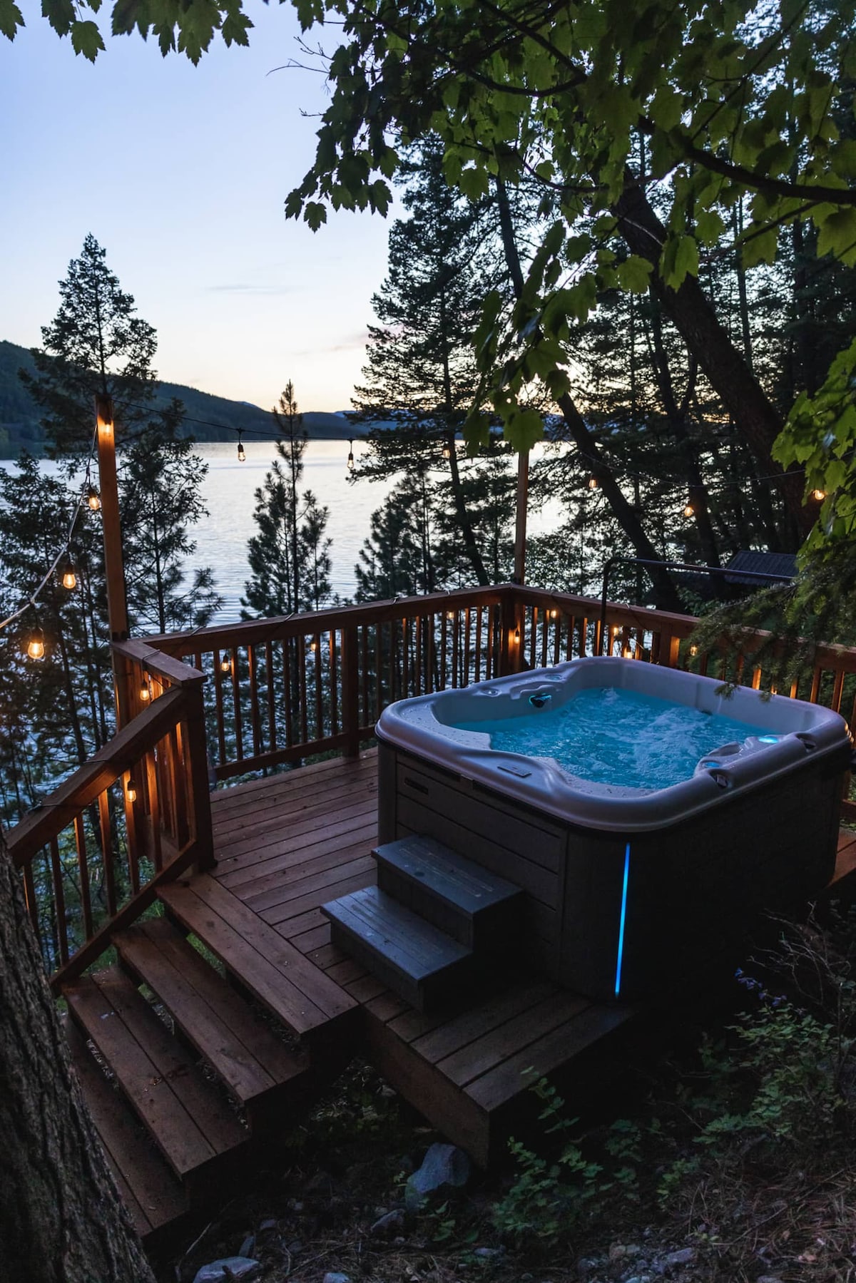 A private hot tub is situated on a wooden deck surrounded by lush trees, providing views of the tranquil lake at twilight. Soft lighting from string lights enhances the serene atmosphere, while the gentle ripples of the water reflect the fading light of the evening sky.