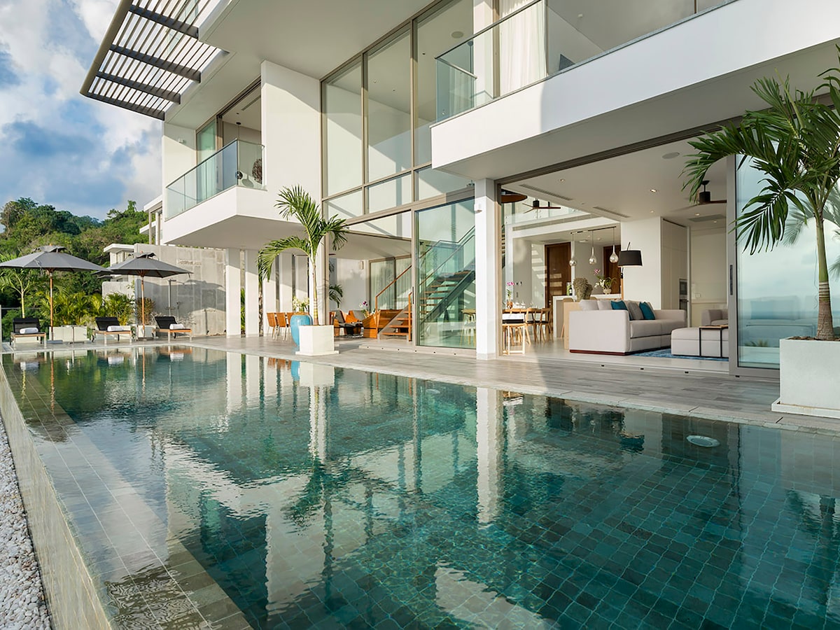 A modern duplex villa features expansive glass walls, showcasing a sleek infinity pool that reflects the surrounding greenery. Sun loungers and umbrellas provide shaded seating by the water, while the open-plan interior is visible through the glass. An elegant stairway leads to the upper level.