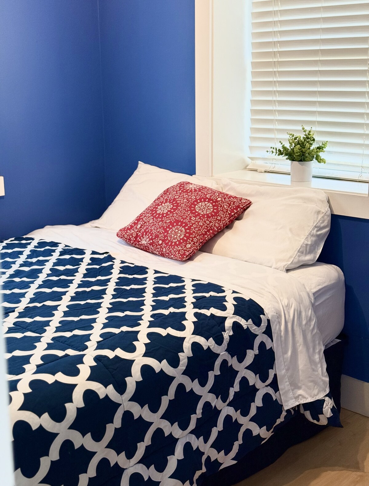 A cozy sleeping area features a compact bed dressed in white linens and a navy patterned quilt. A decorative pillow adds a splash of color, and a small potted plant sits on the windowsill, allowing natural light to brighten the space.