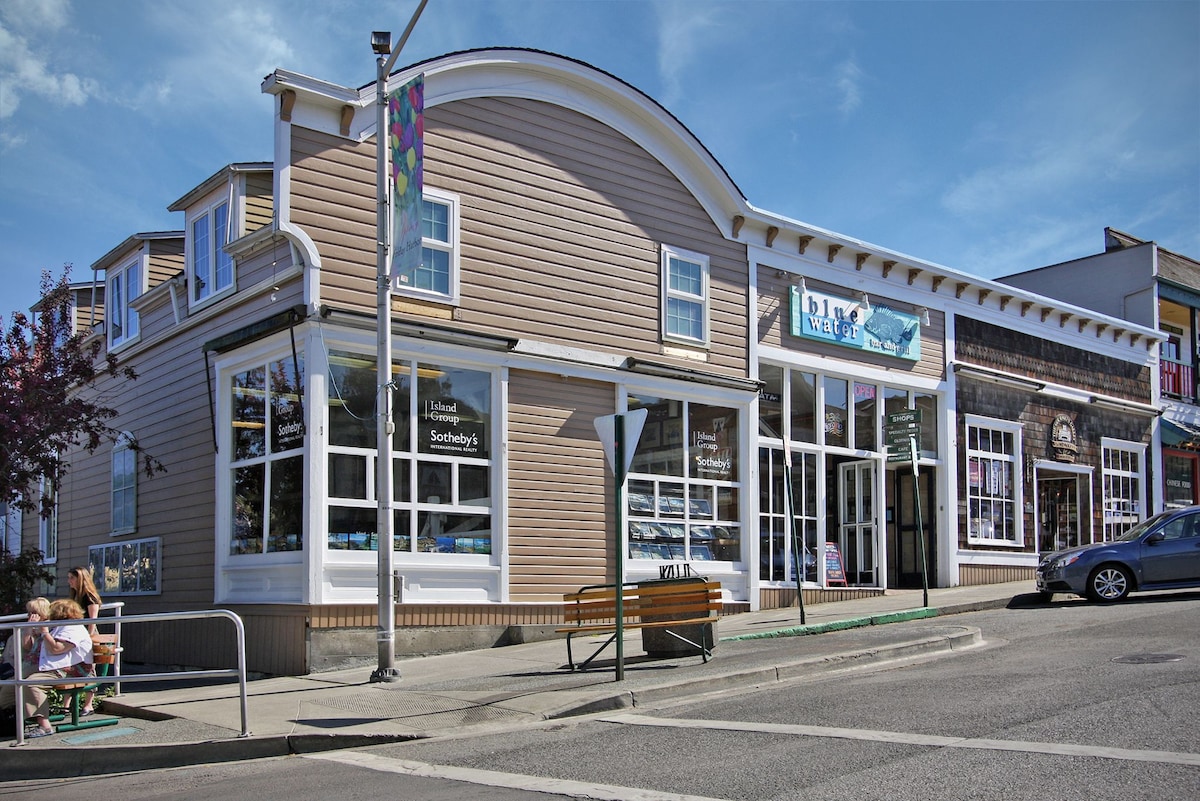 A corner building features a light-colored facade with large windows, allowing ample natural light. Decorative banners are displayed along the outer walls, adding pops of color. A bench is positioned on the sidewalk, enhancing the inviting street view in a bustling area of Friday Harbor.