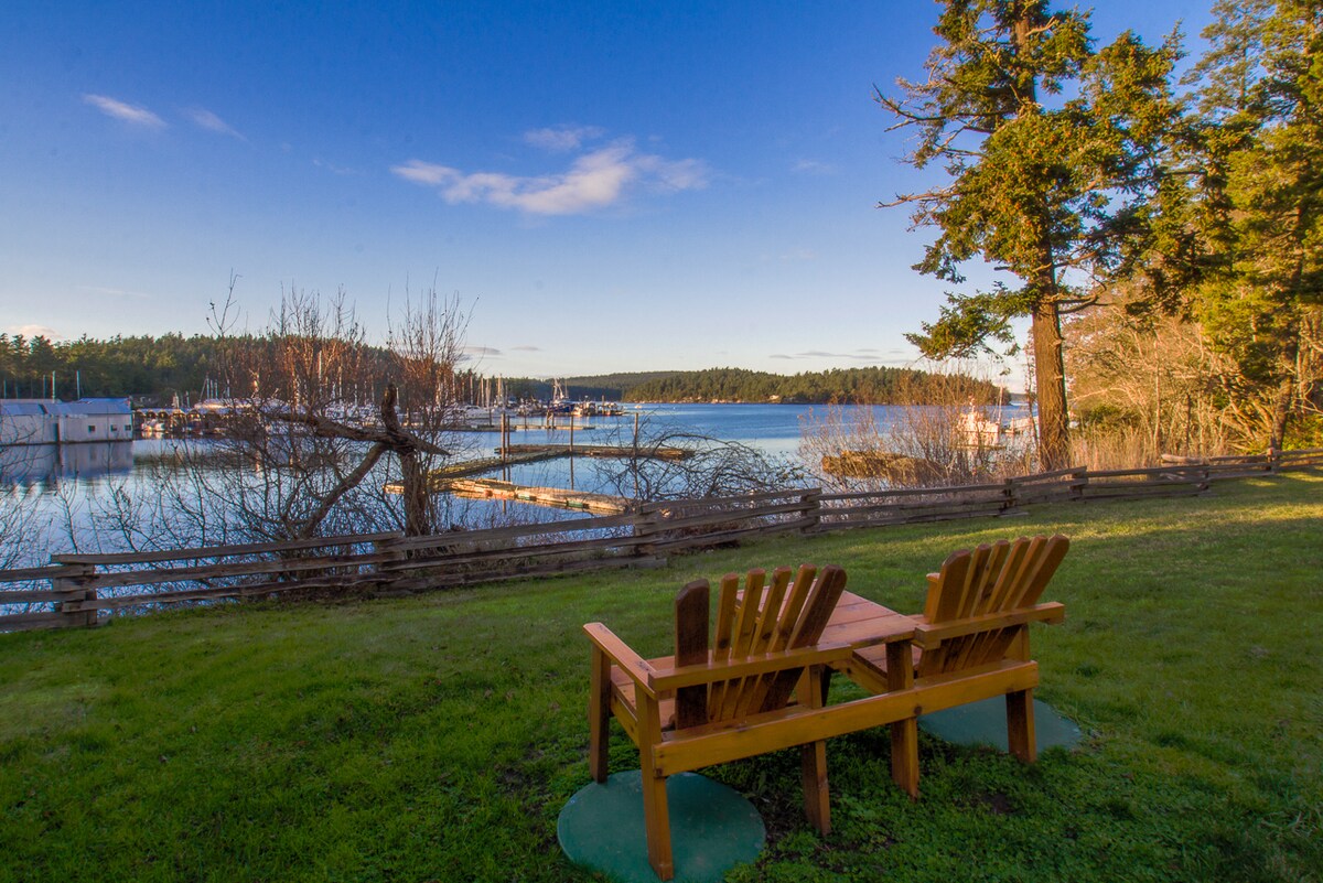 Adirondack chairs sit side by side on a grassy area facing the serene waterfront. The view shows a peaceful marina surrounded by trees, with boats docked in the distance. Clear skies and soft clouds create a calming atmosphere.