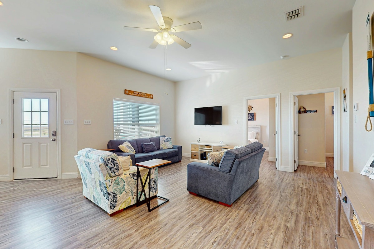 The living area features plush furnishings arranged for comfort and conversation. A 57” smart TV is mounted on a pale wall, while natural light enters through window treatments. The space is designed with an open layout that connects seamlessly to other rooms.