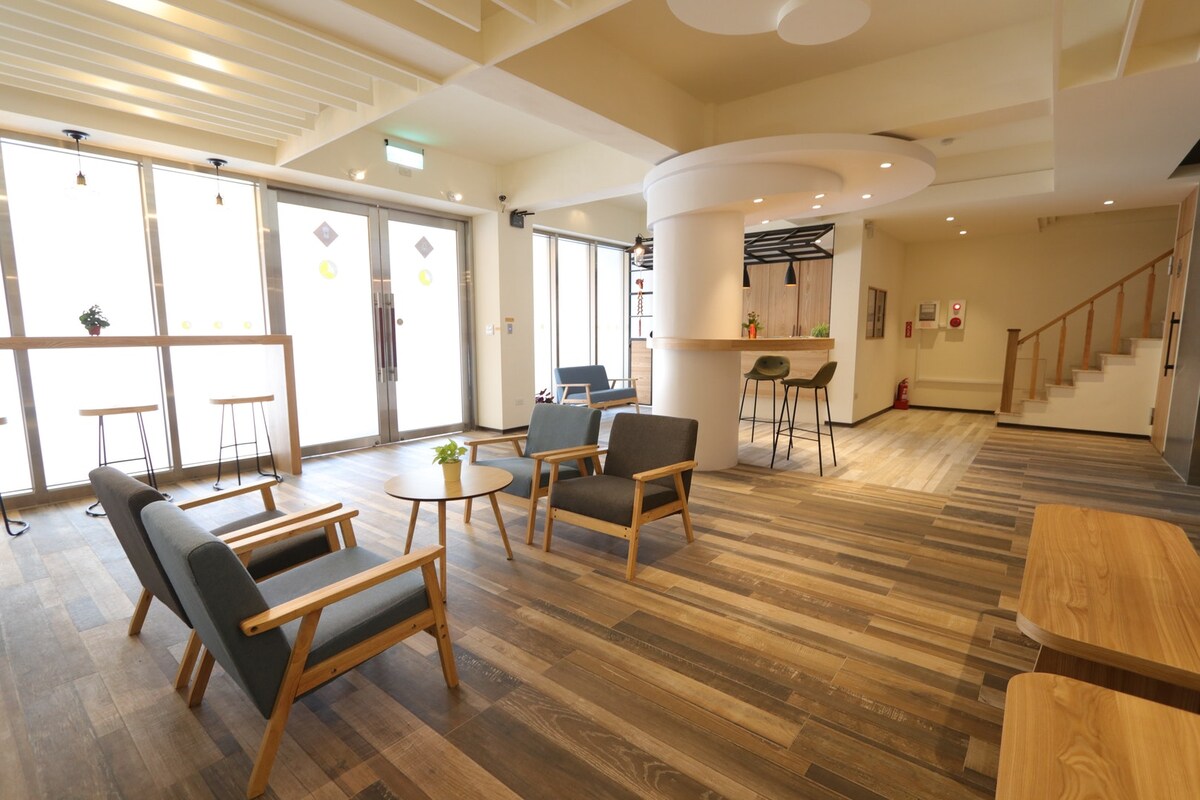 A spacious lobby area features wooden flooring and natural light filtering through large windows. Several wooden tables and comfortable chairs are arranged for relaxation. A modern reception counter is visible in the background, enhancing the welcoming ambiance of the space.
