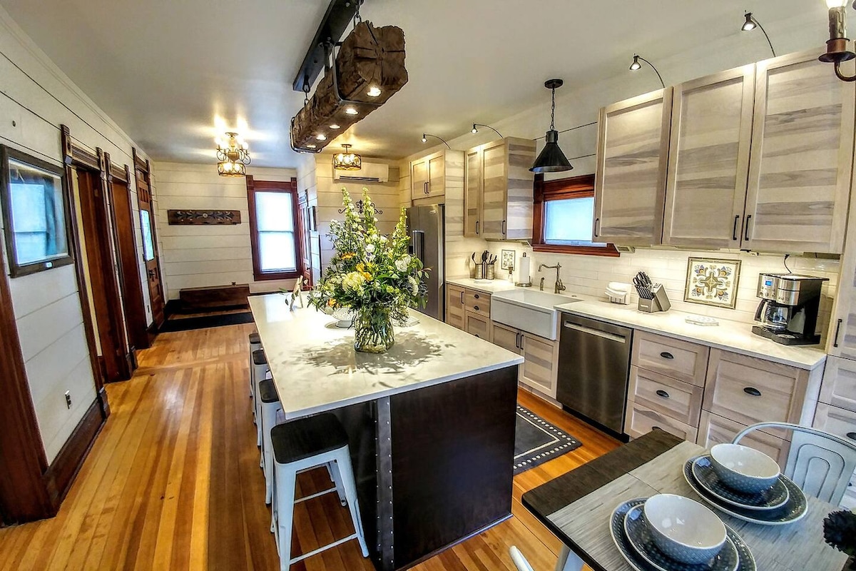 The spacious chef's kitchen, featuring modern appliances and a large island with bar stool seating for four, is well-lit by pendant lights. Natural wood tones highlight the cabinetry, while a decorative flower arrangement adds a touch of freshness to the countertop.