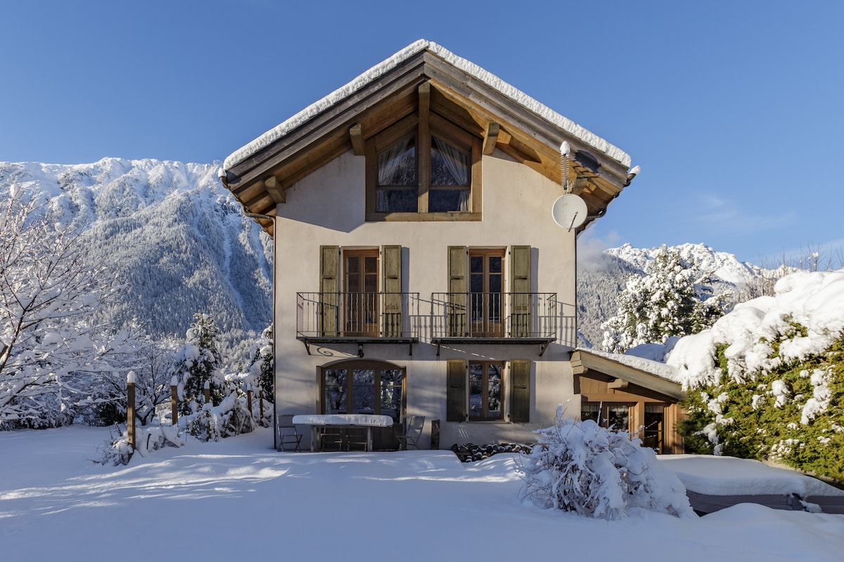 The chalet is framed by a winter landscape, covered in fresh snow. Its traditional architectural style is highlighted by wooden accents and large windows. A balcony extends from the upper floor, while the surrounding trees are adorned with a dusting of snow, creating a serene atmosphere.