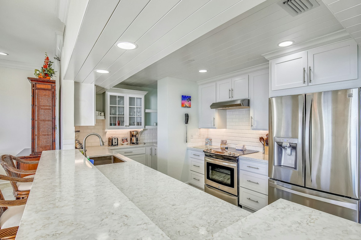 A bright and modern kitchen is showcased, featuring stainless steel appliances and white cabinetry. A large countertop provides ample space for meal preparation. Under-cabinet lighting enhances the work area, while a well-organized pantry is visible in the background.
