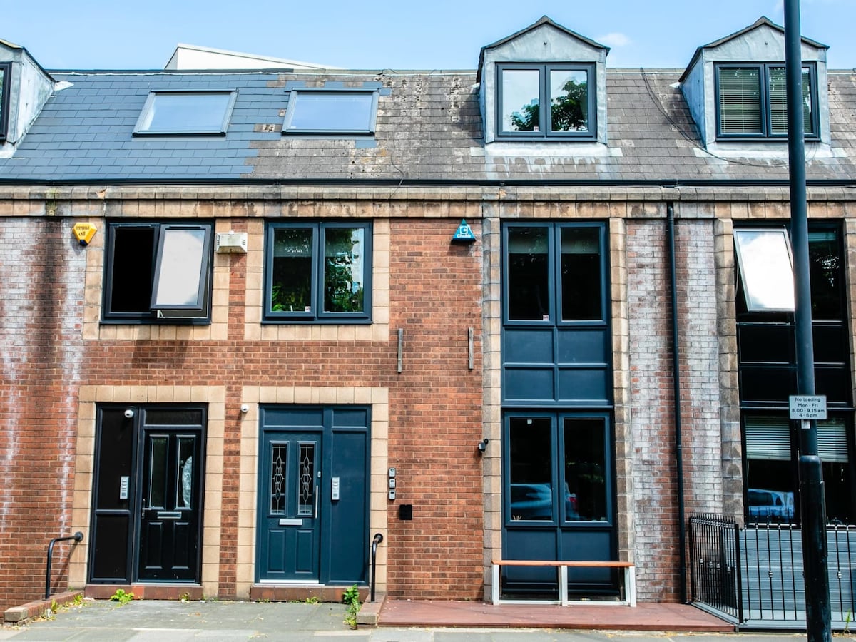 The exterior of a modern townhouse is visible, featuring a blend of brick and grey stone. Large windows allow natural light to enter, while two distinct entrances are positioned at the front. A small bench is located beside the entrance, enhancing the welcoming appearance.