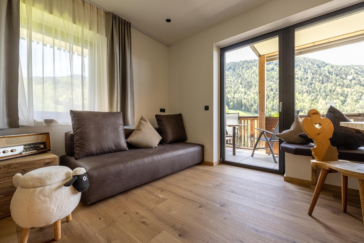 A cozy living area is highlighted by a spacious sofa adorned with decorative pillows. Natural light floods through large sliding glass doors that open to a balcony, revealing a view of green hills. A small wooden table and a playful sheep-shaped stool add charm to the space.