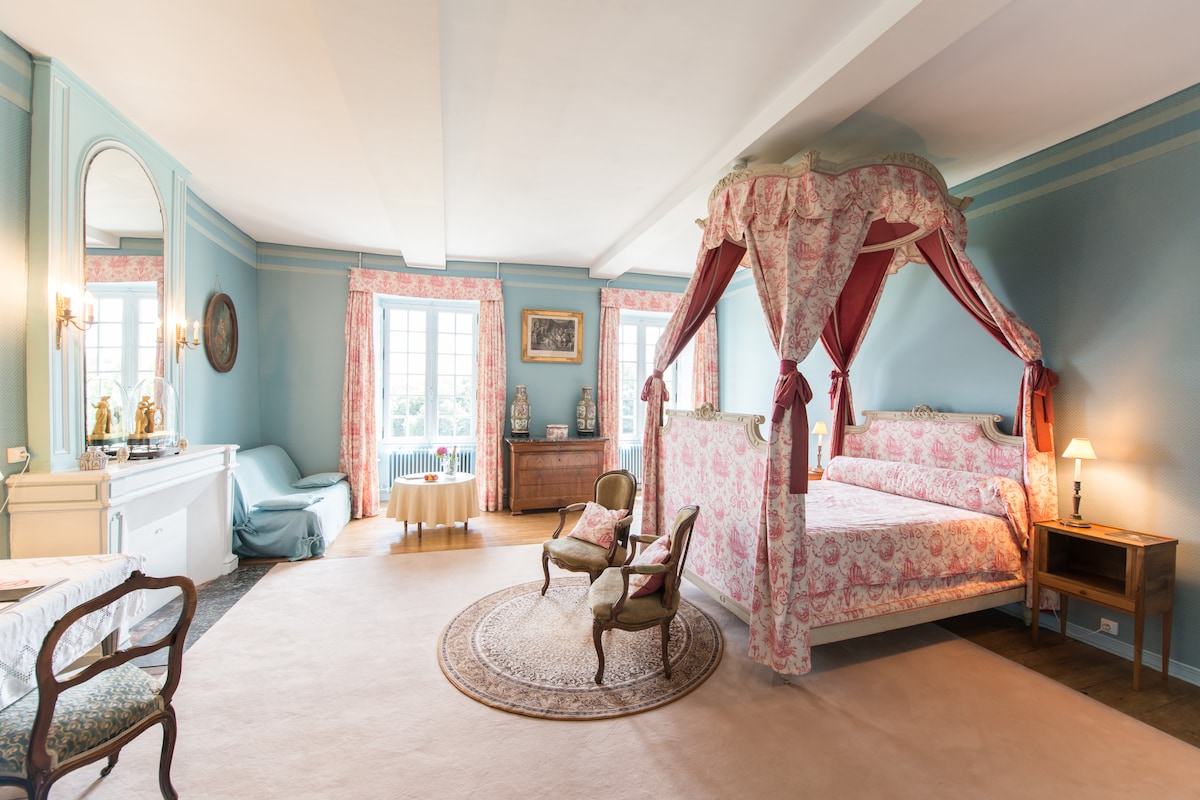 A spacious suite is highlighted by a four-poster bed with a floral canopy. Soft blue walls complement pink floral drapes. A large ornate mirror and traditional furniture, including two elegant chairs, enhance the room, while natural light filters through multiple long windows.