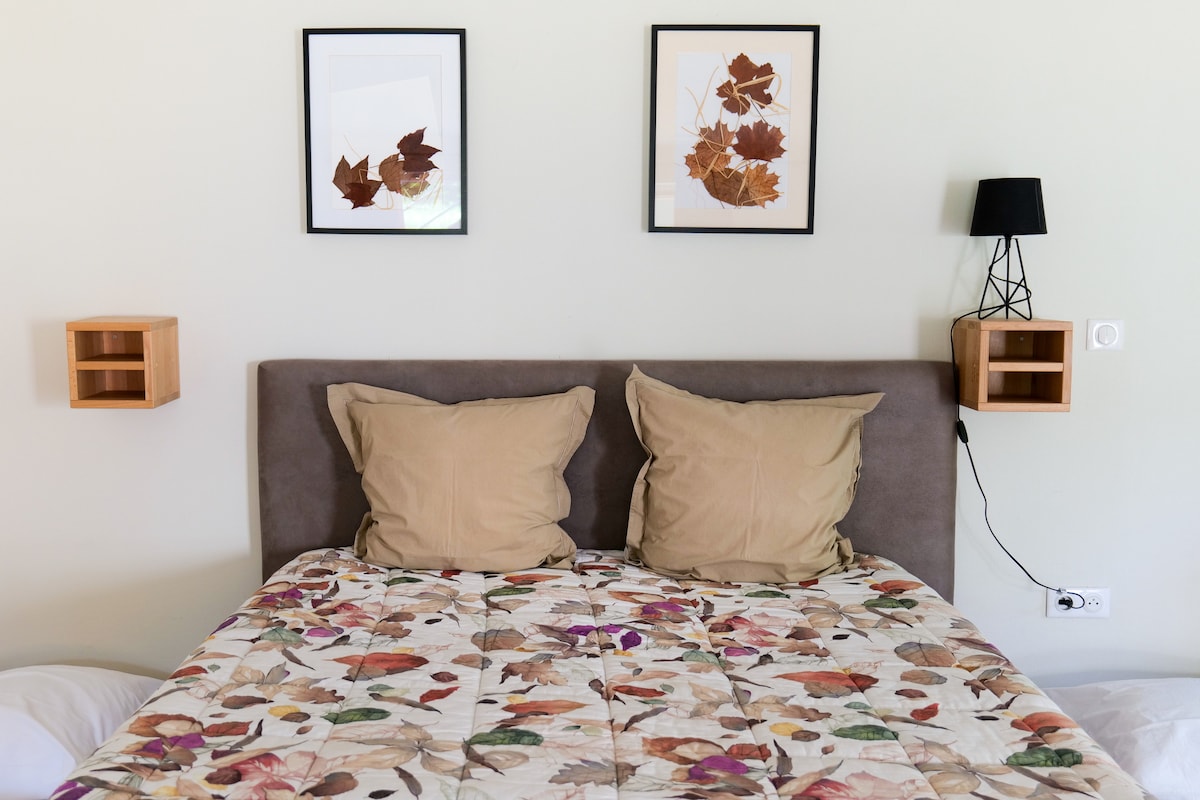 A spacious bed is adorned with a patterned quilt featuring autumn leaves. On the wall above, two framed artworks display dried leaves. Each side of the bed is complemented by a small wooden shelf, while a sleek lamp is mounted on the wall.