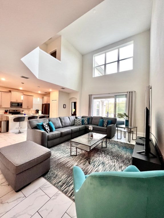 A spacious living area features a large sectional sofa adorned with decorative cushions. A glass coffee table rests on a textured area rug, and natural light floods in through tall windows, casting a warm glow on the open floor plan. A television is positioned for easy viewing.