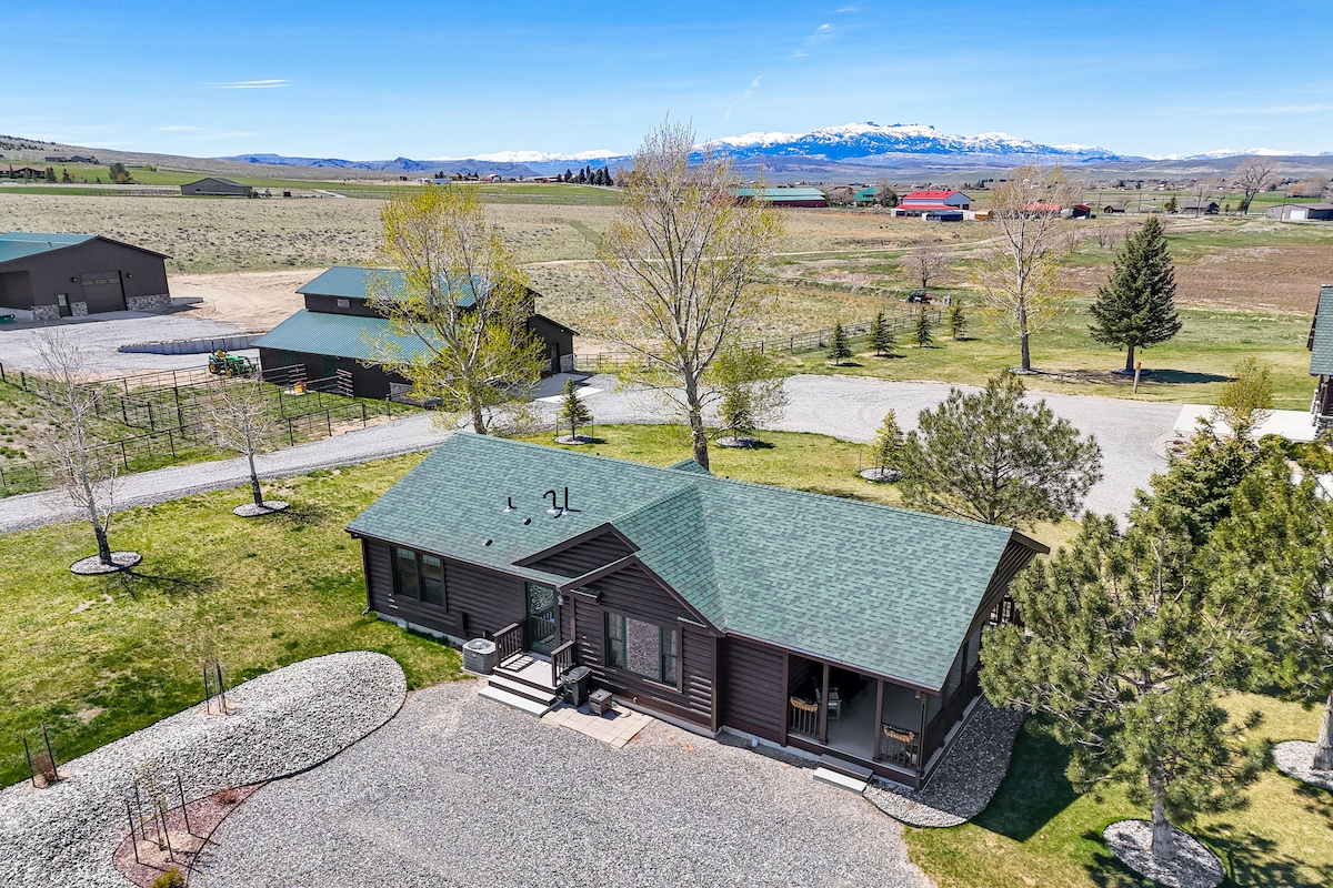 An aerial view showcases a single-story vacation rental featuring a green roof and dark wood siding. Surrounding landscaping includes gravel paths and well-maintained grassy areas. The backdrop reveals expansive ranch land with distant mountains and various structures, highlighting the rural setting.