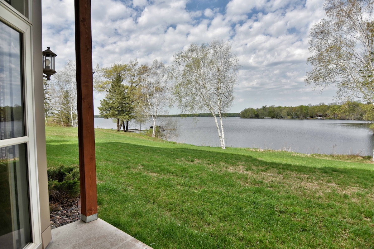 A serene view of Red Cedar Lake can be seen from the patio, showcasing gentle waves and greenery along the shoreline. Soft clouds drift across a blue sky, creating a peaceful outdoor atmosphere framed by a wooden patio structure.