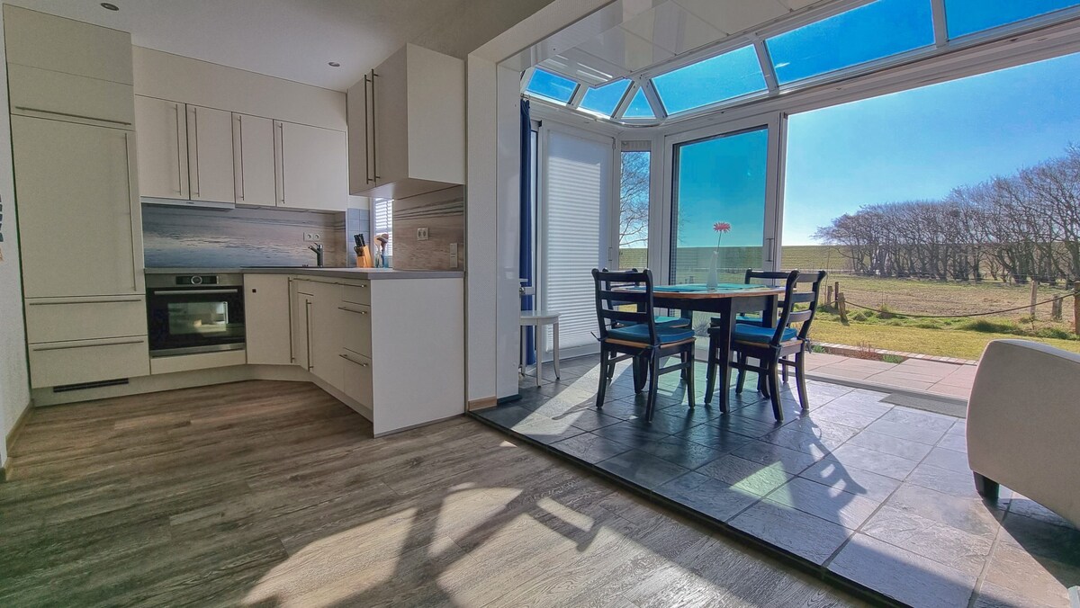 An open-plan kitchen features modern cabinetry and built-in appliances, including an oven. Adjacent is a bright dining area with a round table and four chairs, surrounded by glass walls that provide expansive views of the outdoor landscape and let in ample natural light.