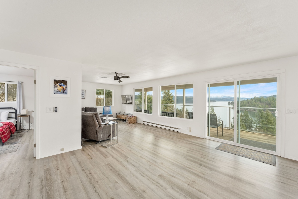 A spacious living area features large picture windows offering views of Lake Coeur d'Alene and natural light throughout. Comfortable seating is arranged around a television, with the open layout extending towards sliding glass doors that lead to an outdoor deck.