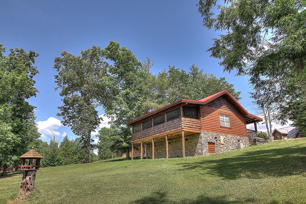 A spacious log cabin is set against a backdrop of lush greenery and clear blue skies. The building features a combination of stone and wood exteriors, with a red-roofed upper section. Surrounding trees and an open lawn contribute to a serene outdoor environment.