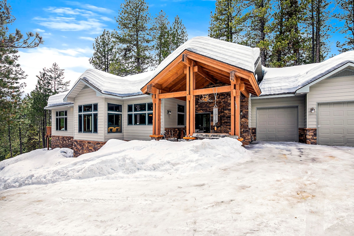 The modern cabin is set against a backdrop of snow-covered ground and tall pines. A spacious entryway features a high, peaked roof supported by wooden beams, and large windows allow for natural light. The driveway leads to a two-car garage on the right.