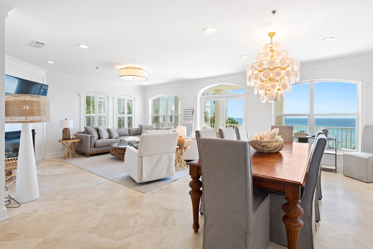 An open living area is illuminated by natural light, featuring a comfortable sectional sofa facing a large TV and a gas fireplace. A dining table with elegant seating accommodates ten. Expansive windows offer views of the ocean, enhancing the inviting atmosphere.
