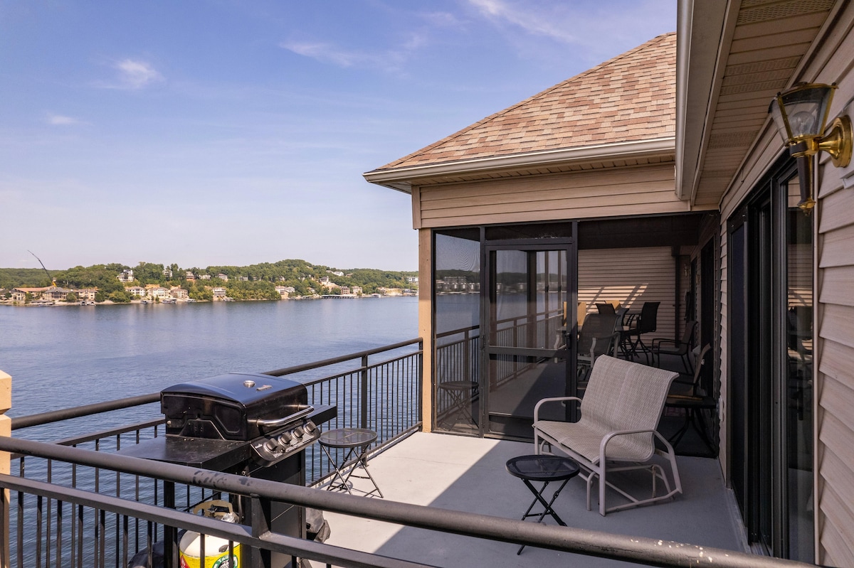 An outdoor balcony presents a stunning view of the lake, featuring a glass railing for unobstructed sightlines. A grill and simple patio furniture, including two chairs and a small table, are positioned to create a relaxing space for guests.