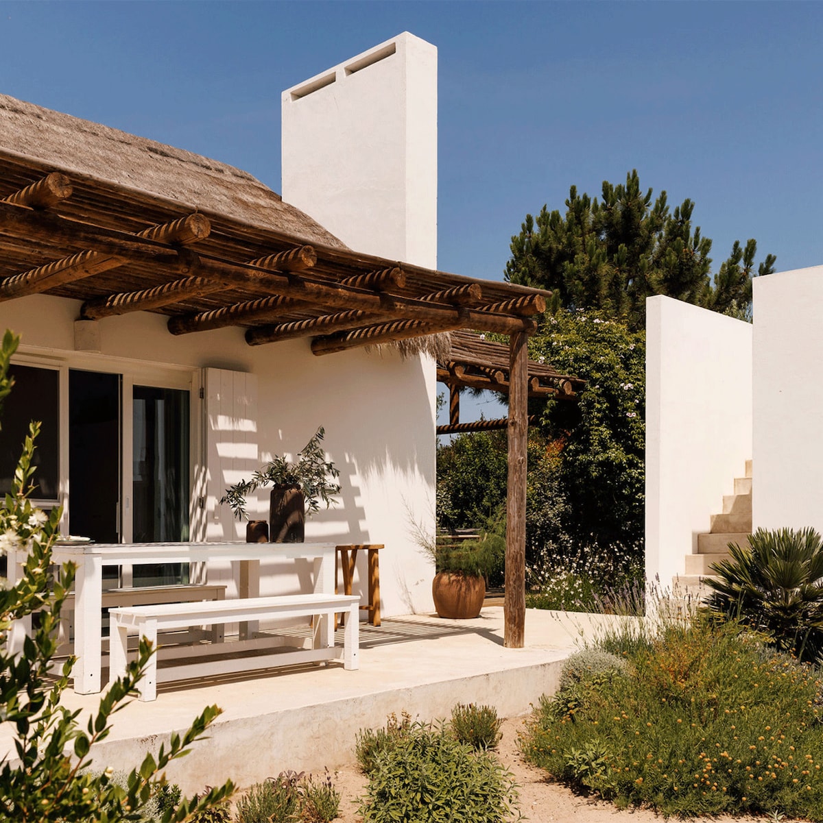 A contemporary white house with a thatched roof is surrounded by a landscaped garden. A wooden pergola supports an outdoor dining area, with a table and chairs positioned on a sunlit terrace. Steps lead to a higher garden level, providing a natural separation.