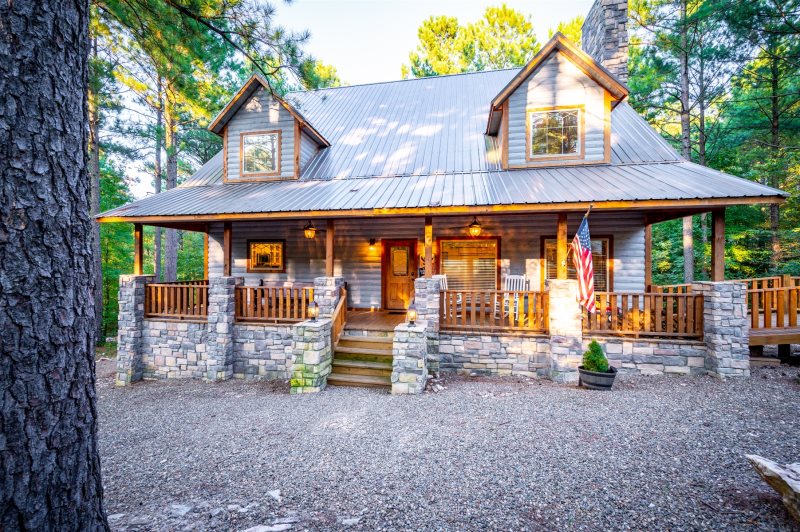 A rustic two-story cabin features a wooden exterior with stone accents. The wraparound porch, framed by tall trees, provides ample seating. Dual dormer windows are positioned above the entrance, and an American flag is displayed prominently. The natural landscape enhances the cabin's inviting appearance.