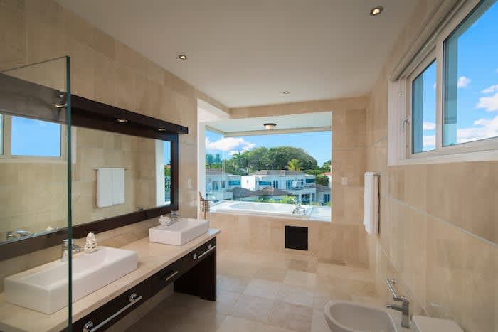 A spacious bathroom is presented with modern features, including dual sinks on a dark wooden vanity. Natural light enters through large windows, revealing views of the surrounding villa area. Neutral-toned stone tiles cover the floor and walls, enhancing the contemporary design.
