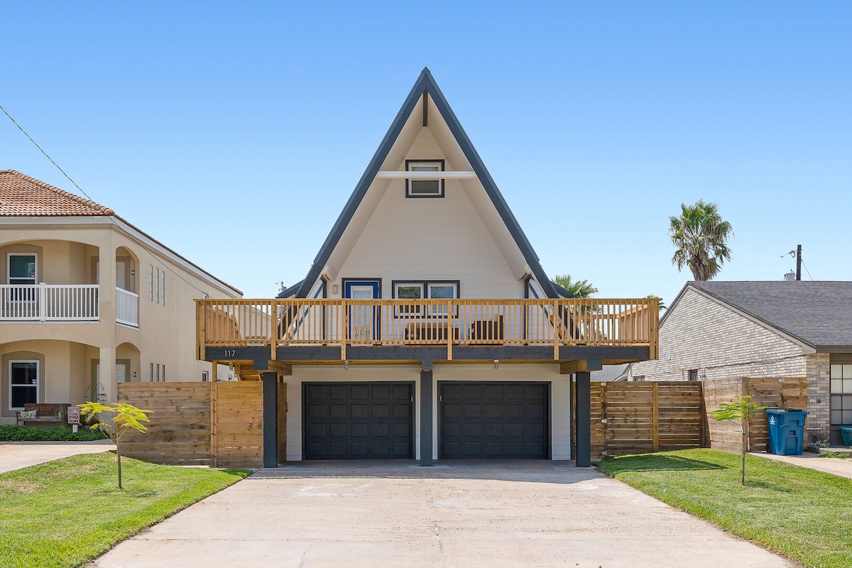 A two-story A-frame house is shown with a spacious wooden deck at the front, supported by wooden posts. The property features dual garage doors, and green landscape elements including young trees are present on either side of the driveway.