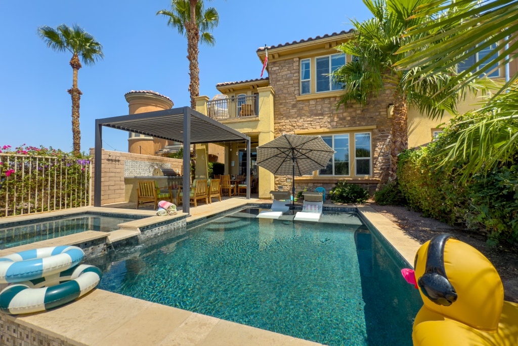 A vibrant outdoor space features a shimmering pool, flanked by palm trees and colorful landscaping. A shaded seating area with an umbrella offers comfort, while lounge chairs invite relaxation. Floating pool toys rest on the water's surface, enhancing the inviting ambiance of the luxurious retreat.