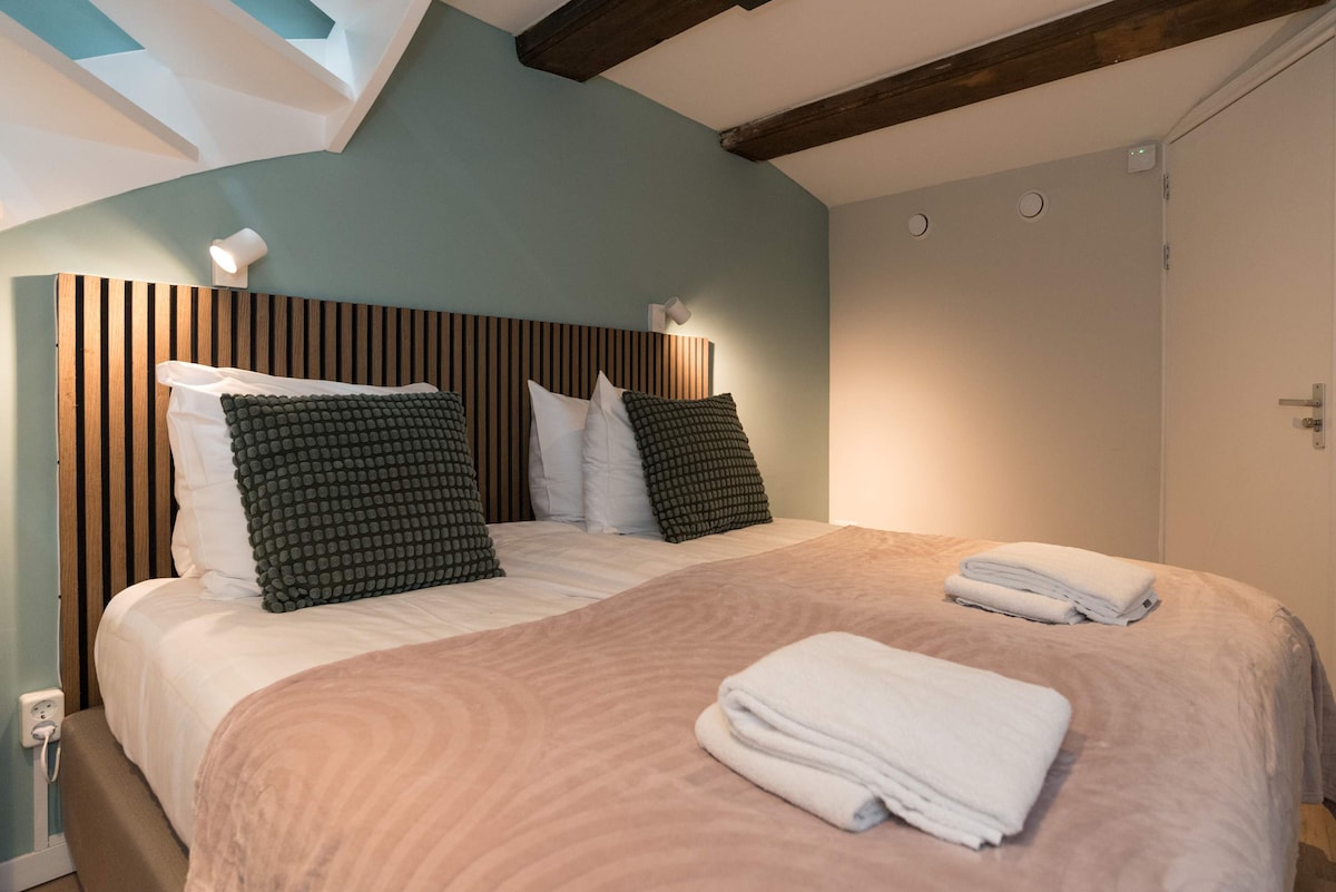 A well-arranged sleeping area features two spacious beds, each adorned with textured pillows and a soft blanket. Neutral wall colors are complemented by wooden beams overhead, while soft lighting creates a calm ambiance. Fresh towels are neatly placed at the foot of one bed.