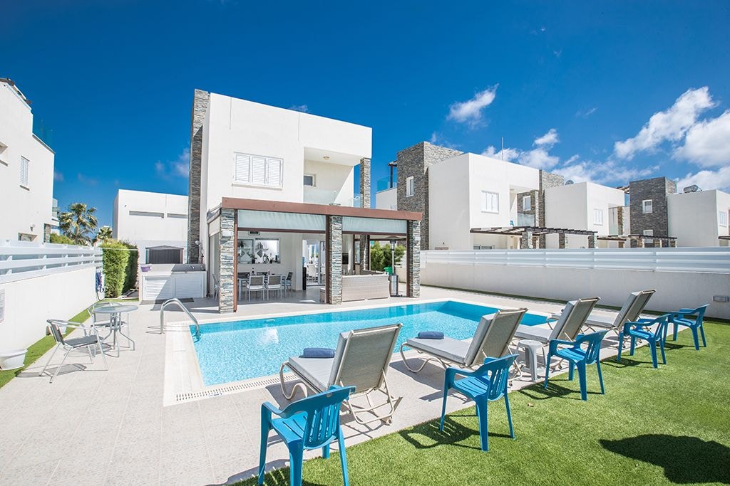 The outdoor area features a private swimming pool surrounded by comfortable sun loungers. A covered seating area provides shade and overlooks the pool, while the well-maintained garden includes AstroTurf. The modern villa's clean lines and spacious design are highlighted against a clear blue sky.