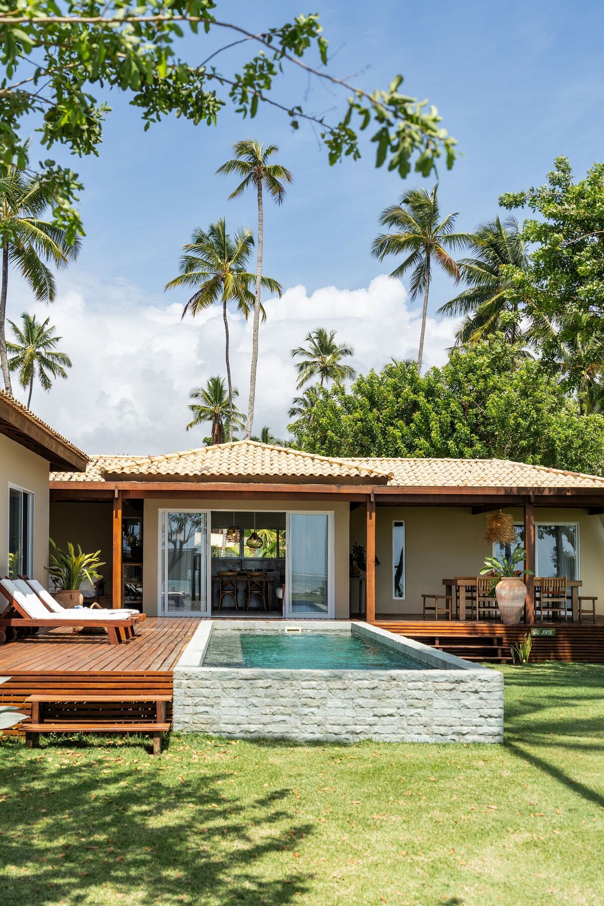 The image captures a tropical residence featuring a swimming pool surrounded by a wooden deck. Palm trees are visible in the background, and an outdoor dining area is set beneath a roofed porch, seamlessly integrating indoor and outdoor living spaces.