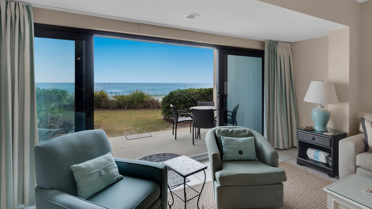 The living area features soft furnishings in calming hues, with two comfortable chairs facing an expansive sliding glass door that opens to a private patio. The view extends to the Gulf, framed by lush greenery, creating an inviting connection to the outdoor space.