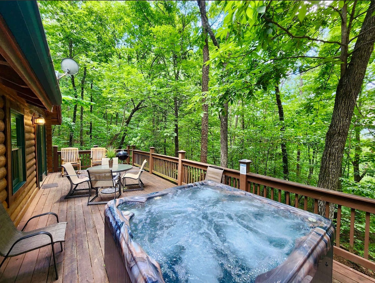 A private hot tub is situated on a spacious wooden deck surrounded by lush greenery. Comfortable seating, including chairs and a table, invites relaxation. The cabin blends harmoniously with the natural setting, featuring a rustic log exterior and tall trees that provide a serene atmosphere.