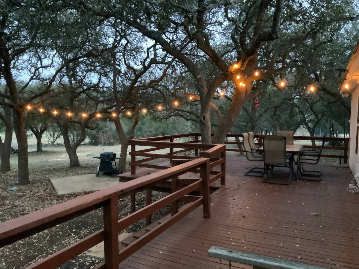 An outdoor deck is framed by mature trees adorned with string lights, providing a soft glow. A gas grill is positioned nearby, and a dining table with chairs invites outdoor meals. The surrounding area is filled with natural foliage and a spacious, open atmosphere.