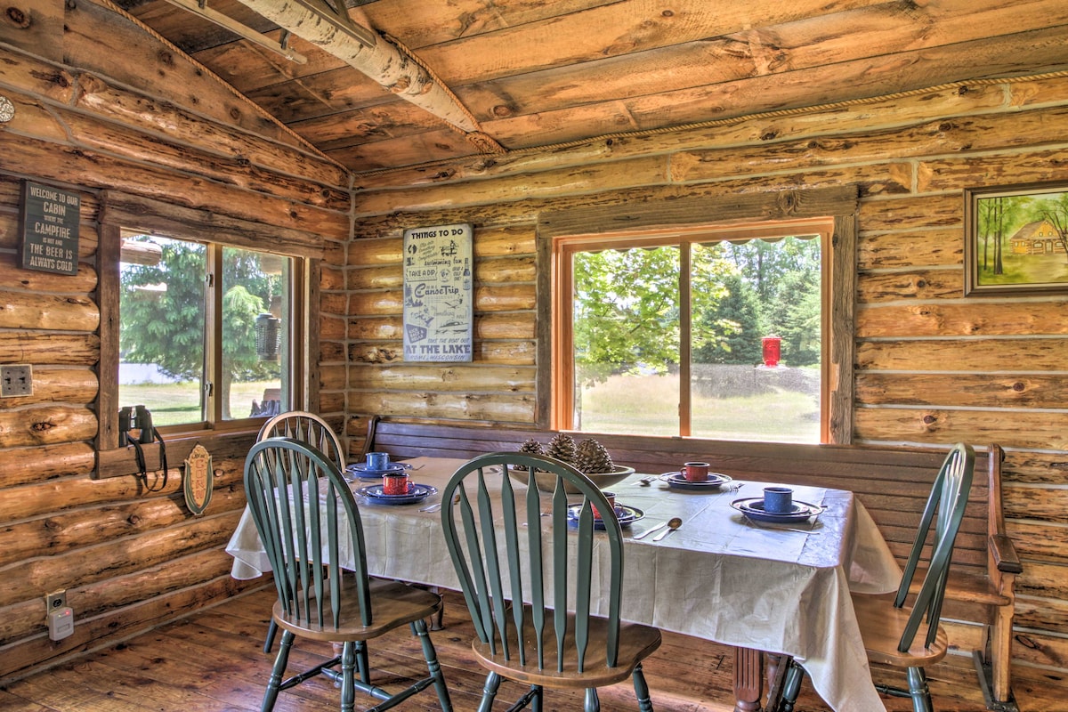 A charming dining area features a wooden table set with blue dishes and utensils. The surrounding log walls add rustic appeal, while large windows provide natural light and views of the outdoors, enhancing the inviting atmosphere of the cabin.