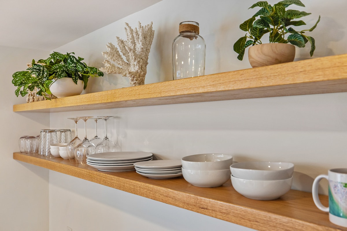 Open shelving displays an assortment of tableware, including glassware, plates, and bowls. Potted plants add a touch of greenery above the polished wooden shelves, contributing to a clean and organized look in the kitchen area.