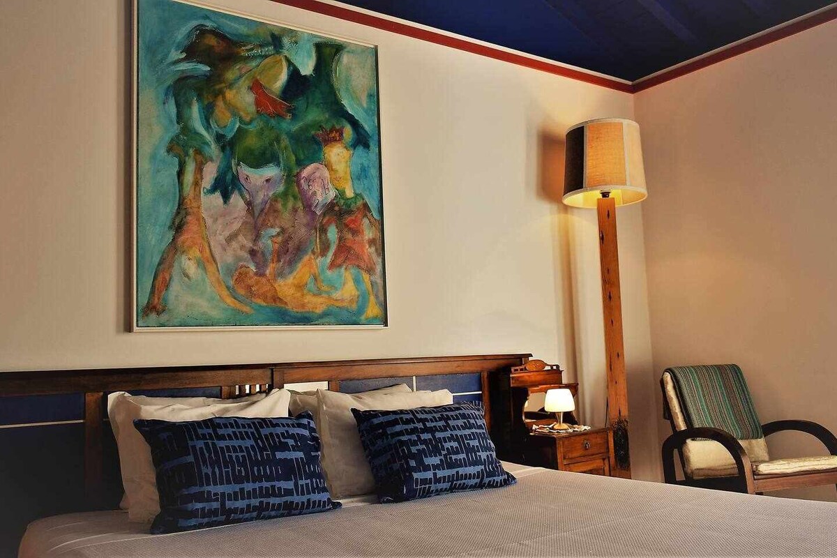 A comfortable bedroom is presented with a spacious bed dressed in crisp linens and accented by decorative pillows. A colorful artwork adorns the wall above the bed. A side table holds a lamp, complementing a cozy armchair and a chaise positioned nearby.