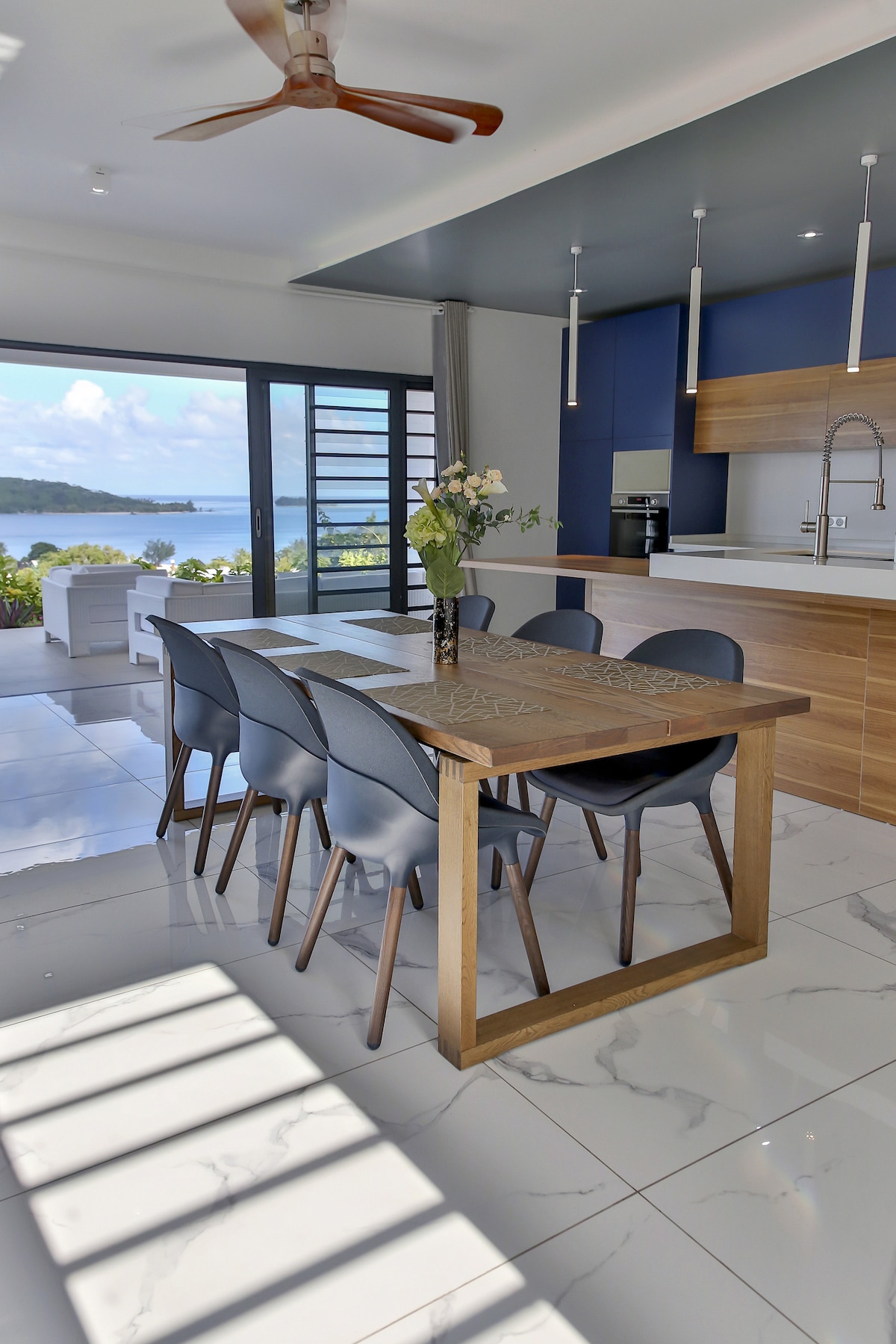 A dining area is presented with a wooden table surrounded by six upholstered chairs. Large glass doors provide a view of the lagoon, while a modern kitchen with sleek cabinetry is visible in the background. Sunlight creates a pattern on the glossy tile floor.
