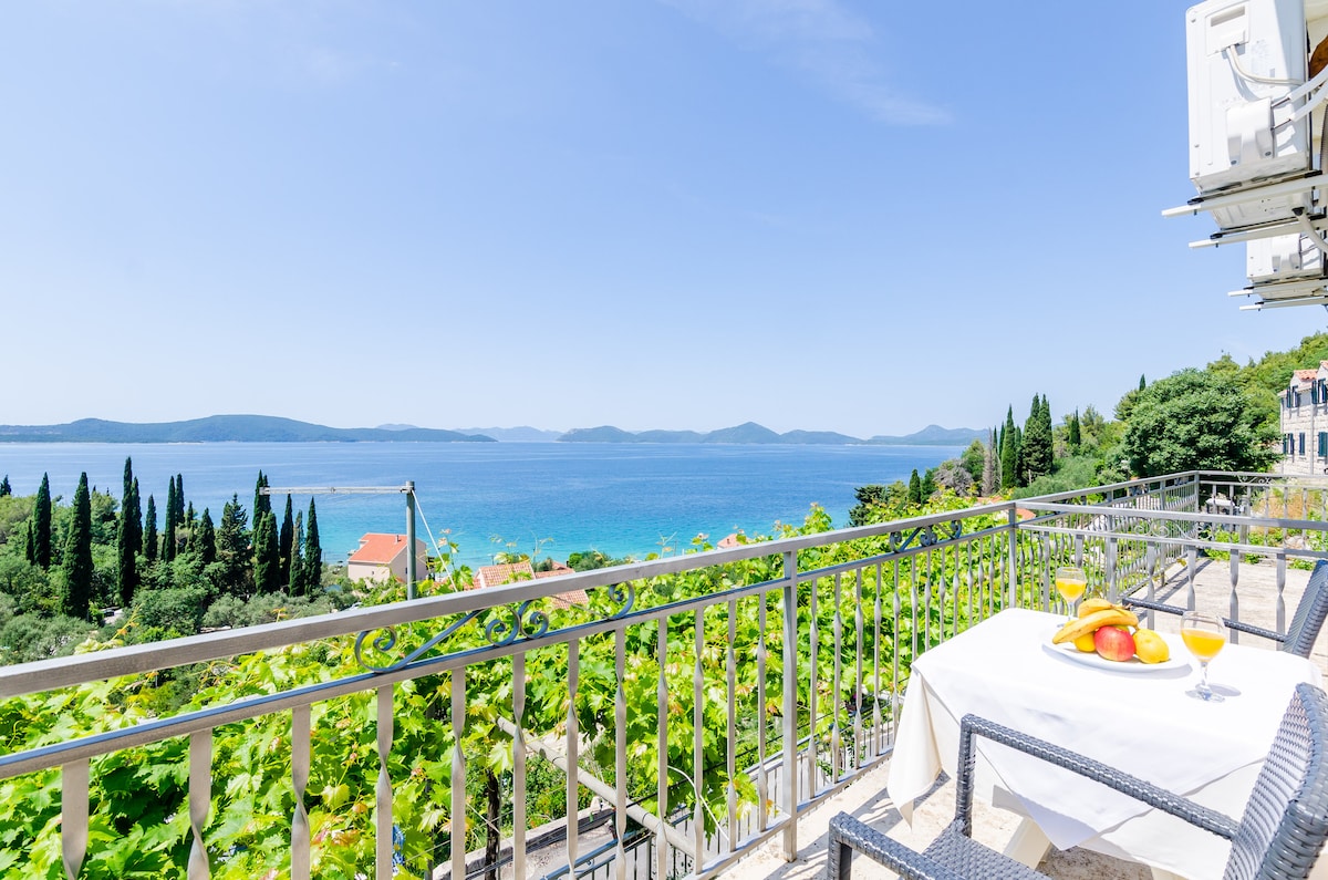 A spacious balcony is presented with outdoor furniture, offering panoramic views of the sea and distant islands. A table is set with a bowl of fruit and two glasses of orange juice, surrounded by lush greenery that enhances the tranquil setting.