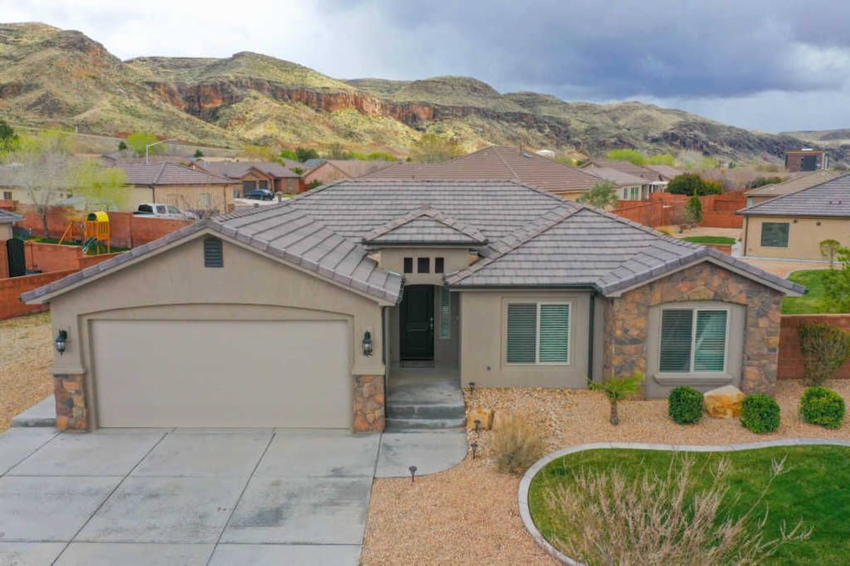 A single-story home is depicted in a spacious lot, featuring a stone-accented facade. The driveway provides ample space for parking. Surrounding mountains are visible in the background, while landscaped areas include gravel and a small lawn, contributing to the home's inviting curb appeal.